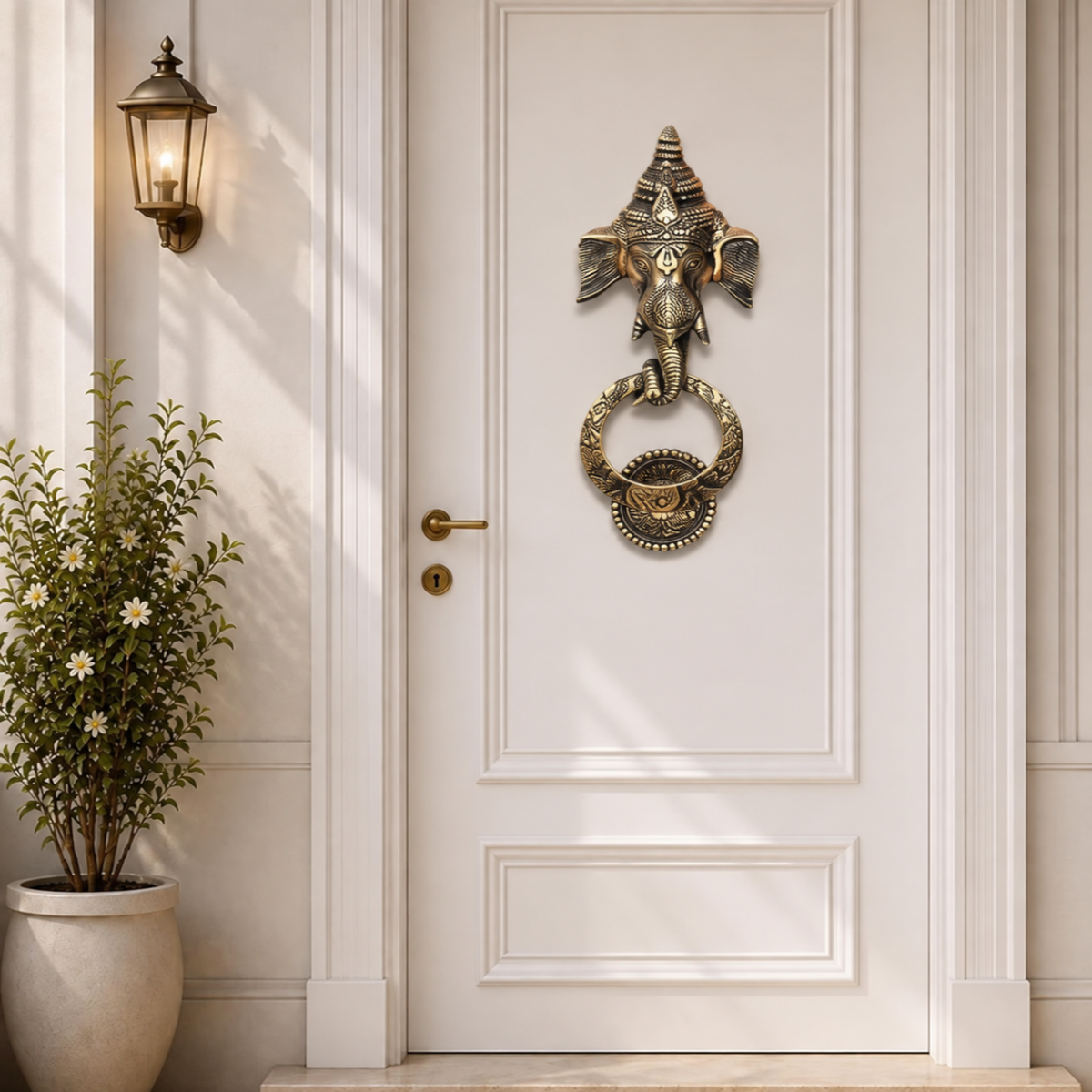 An bronze Ganesha door knocker mounted on a white paneled door, with a matching brass doorknob and a wall-mounted lantern light to the left, alongside a potted plant.