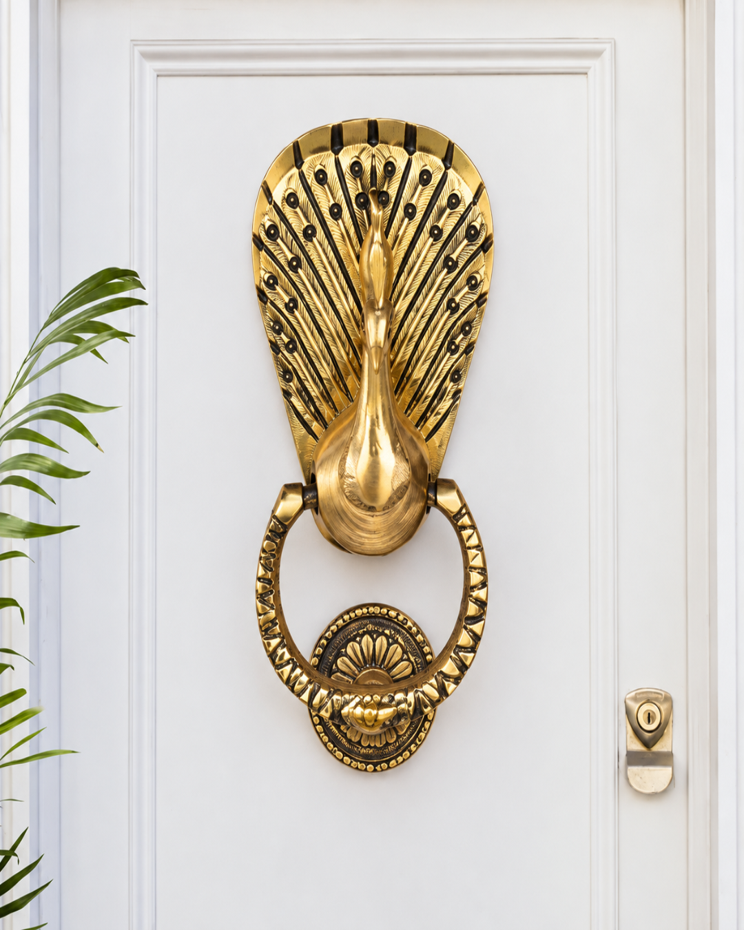 An ornate gold peacock door knocker mounted on a white paneled door, with a small green plant leaf visible on the left and a brass keyhole cover on the right.