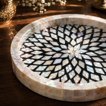 A round mother-of-pearl tray with a geometric fan pattern in iridescent white, beige, and soft purple tones, set on a dark wooden table.