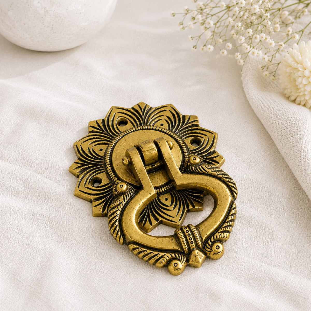 An ornate antique-style brass door knocker with a sunburst backplate, displayed on a textured white fabric with decorative dried flowers and a white stone vase in the background.