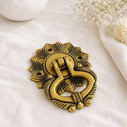 An ornate antique-style brass door knocker with a sunburst backplate, displayed on a textured white fabric with decorative dried flowers and a white stone vase in the background.