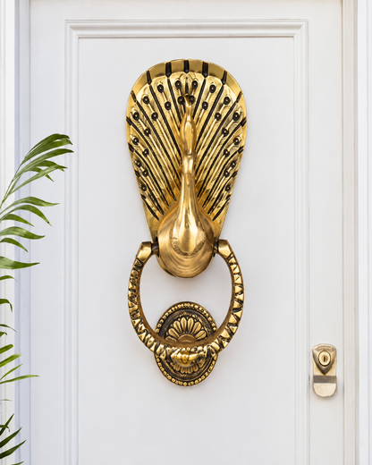 An ornate gold peacock door knocker mounted on a white paneled door, with a small green plant leaf visible on the left and a brass keyhole cover on the right.