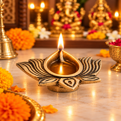A gold-toned lotus-shaped oil lamp with a lit wick and flame, placed on a marble surface in a decorated festive setting with marigold flowers and blurred brass ornaments in the background.