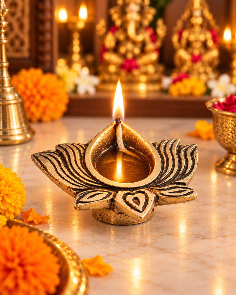 A gold-toned lotus-shaped oil lamp with a lit wick and flame, placed on a marble surface in a decorated festive setting with marigold flowers and blurred brass ornaments in the background.