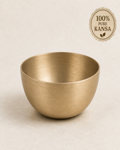 A brushed brass bowl with a matte finish, shown on a light beige textured background with a circular '100% Pure Kansa' badge in the top right corner.