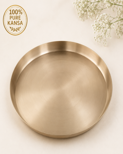A round brushed brass serving tray with a shallow rim, shown on a light cream background with baby's breath flowers in the top right corner and a circular '100% Pure Kansa' brand badge in the top left.