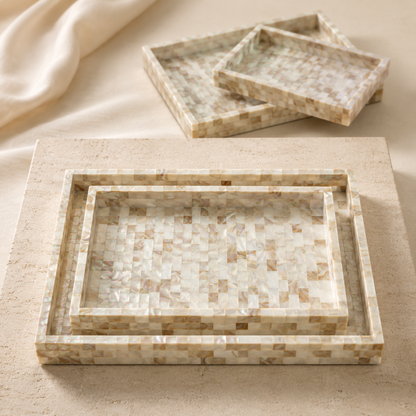 Three nested rectangular trays with a mosaic shell-like pattern in cream, beige, and light brown tones, arranged on a matching textured surface with a folded cream fabric in the background.