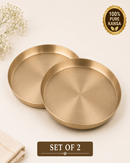 Two round brushed brass Kansa plates shown on a light cream background with decorative baby's breath flowers and a folded cloth. A gold seal reads '100% Pure Kansa' and a label at the bottom says 'SET OF 2'.